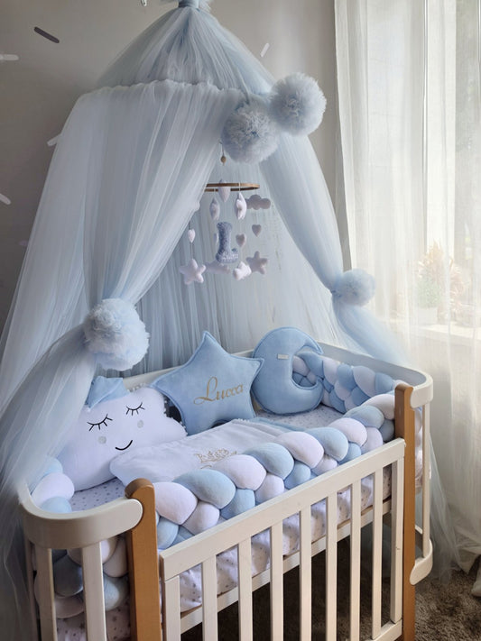 A light blue and white cloud-themed baby canopy bed set with a matching cloud-shaped pillow, plush cloud cushions, and a hanging mobile, styled in a softly lit nursery next to a window with sheer curtains.