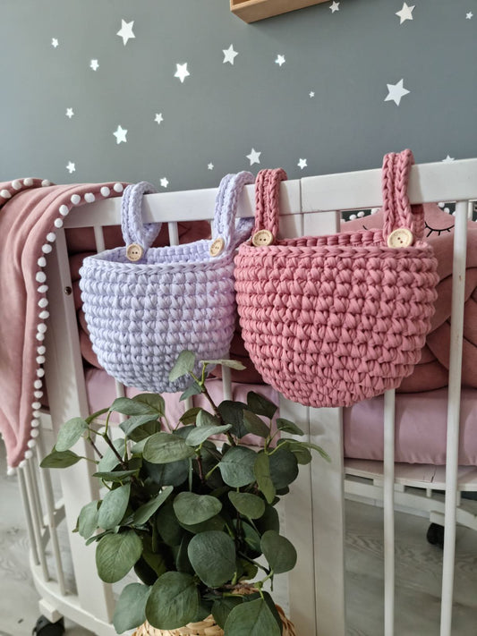 Two crocheted baskets, one pink and one purple, hanging on a crib with a plant in the foreground.