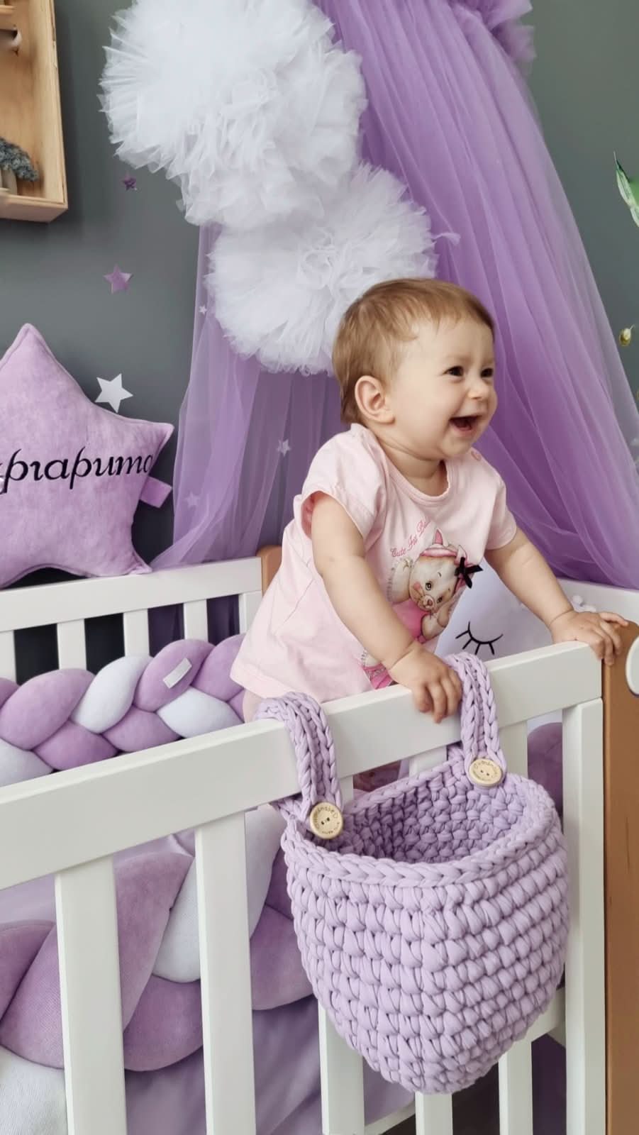 Child in a crib with a purple basket, surrounded by purple decor in a nursery.