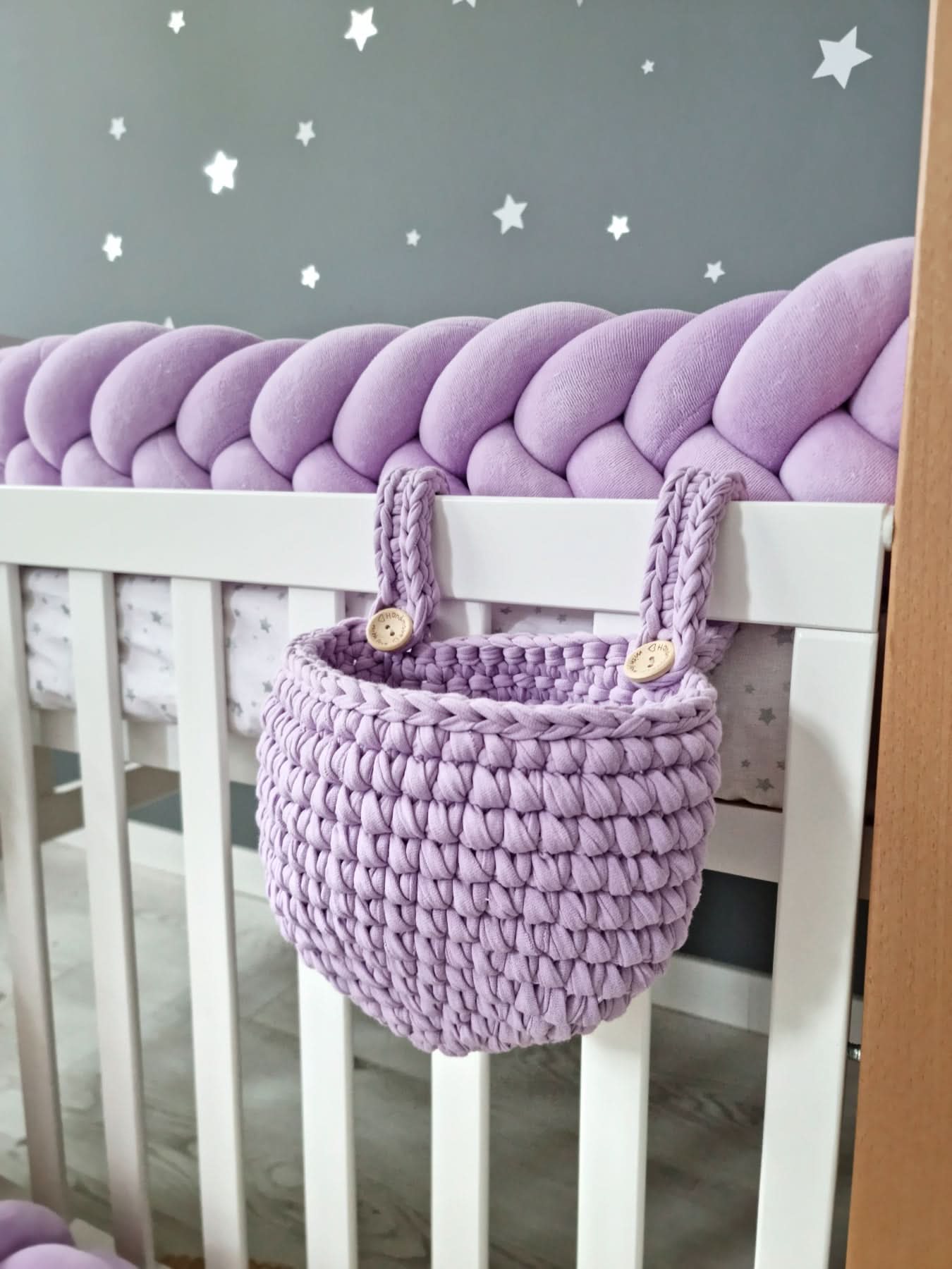 Crib with a purple knitted bumper and matching basket against a gray wall with star decals.