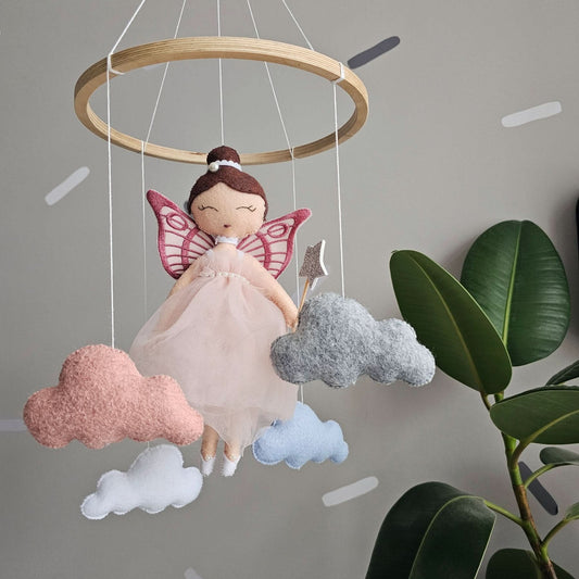 Felt fairy doll with pink wings hanging from a wooden ring against a gray background with plants.