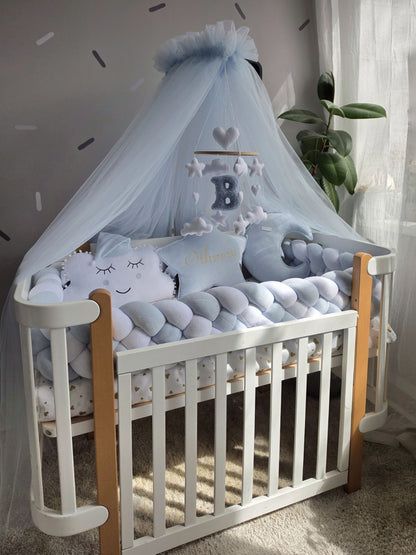 A white wooden baby crib with natural wood rails, styled with a light blue tulle canopy, a plush cloud pillow, a blue letter B mobile, and a matching blue crescent moon pillow in a softly lit nursery.