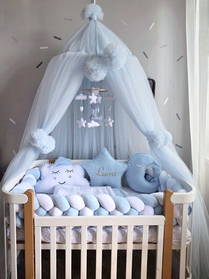 A light blue baby canopy with fluffy pom-poms and a matching star-and-cloud mobile hanging over a wooden crib. The crib is styled with a cloud-shaped pillow, a crescent moon cushion, and a blue star pillow embroidered with the name 'Luca'.
