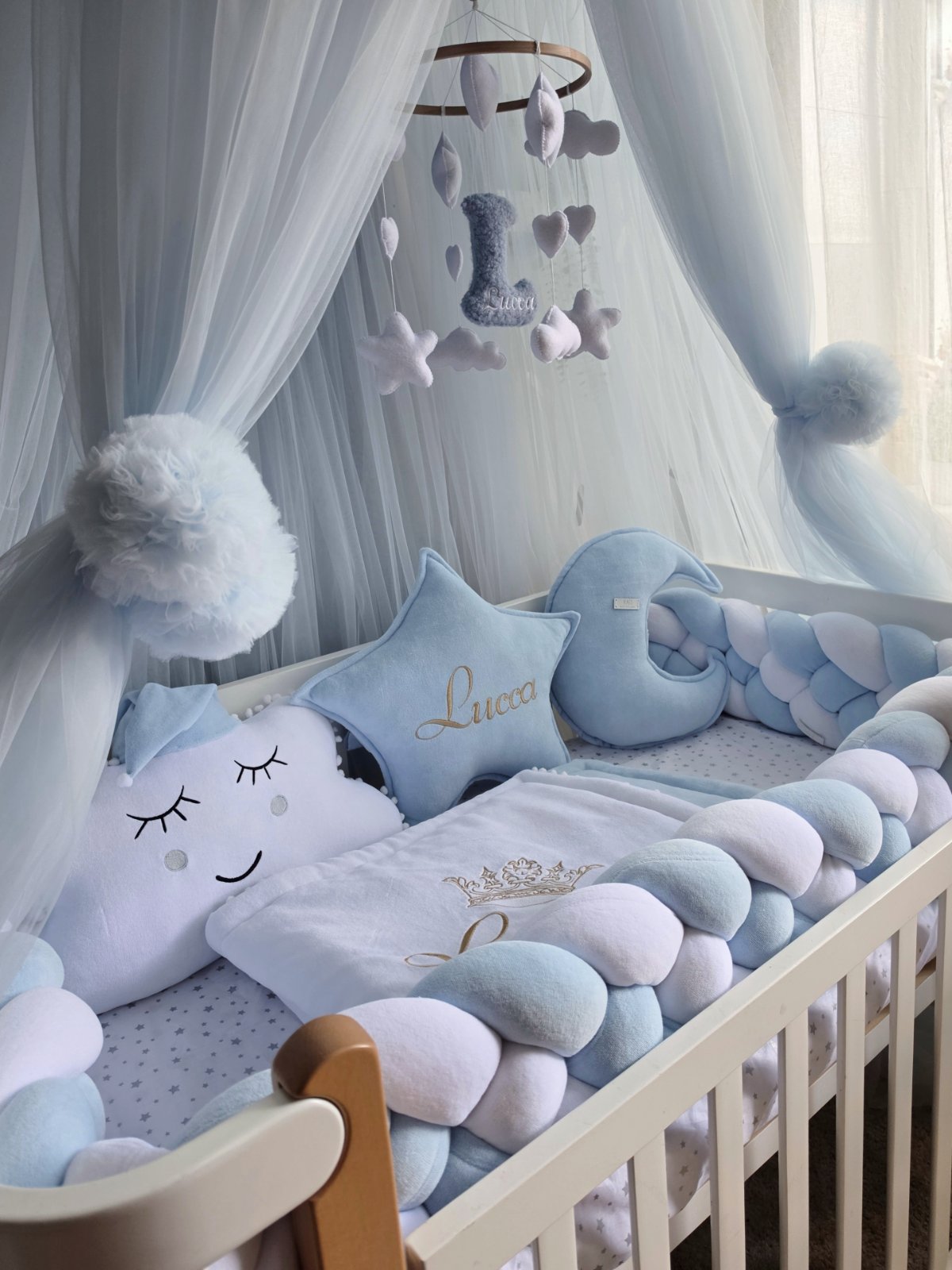 A light blue and white baby crib setup with a sheer canopy, plush cloud pillows, a star-shaped pillow embroidered with the name 'Luca', a crescent moon pillow, and a matching cloud pillow guard.