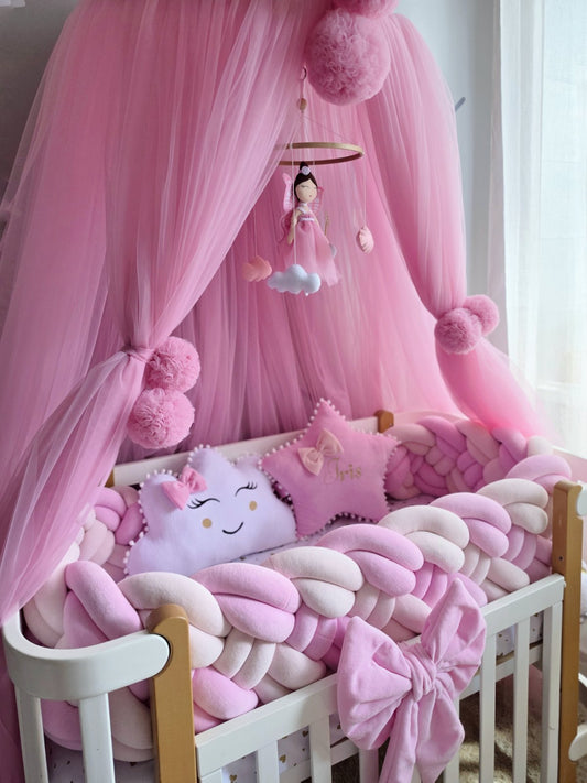 A pink baby crib setup with a white wooden crib, chunky pink and white knit bumper pads, a pink bow, a plush star pillow, a smiling cloud pillow, and a large pink tulle canopy with pom-poms and a hanging mobile.