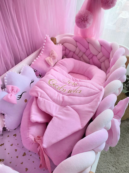 A pink baby bedding set arranged in a crib, featuring a plush pink bassinet cushion, matching pink blanket with gold embroidered text, pink pom-pom pillows, and a lavender star-shaped pillow with a pink bow.