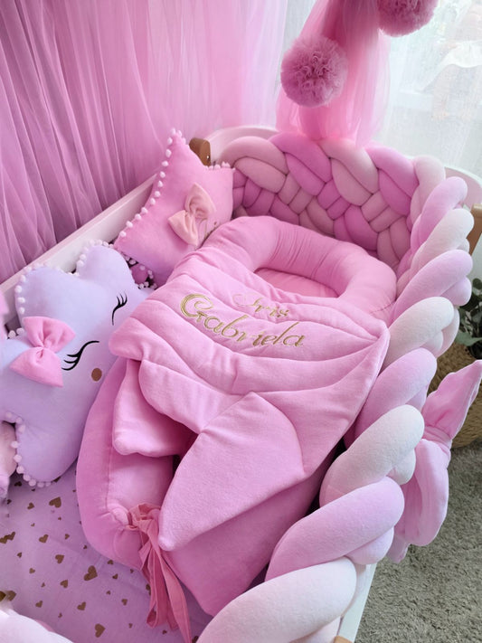 A pink baby bedding set arranged in a crib, featuring a plush pink bassinet cushion, matching pink blanket with gold embroidered text, pink pom-pom pillows, and a lavender star-shaped pillow with a pink bow.
