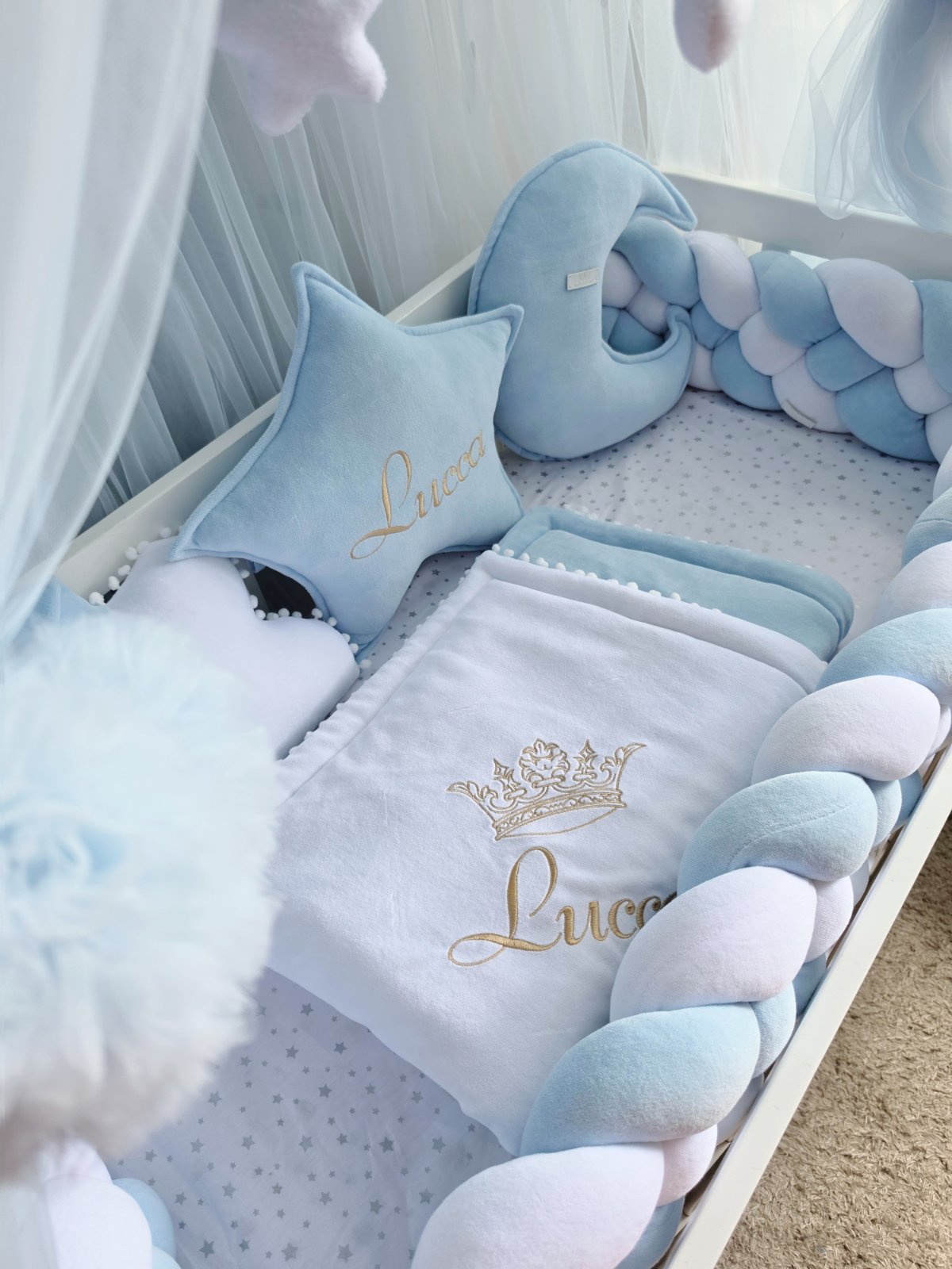 A light blue and white baby bedding set arranged in a crib, featuring a plush blanket with a gold embroidered crown and the name 'Lucas', a matching star pillow, a crescent moon pillow, and a braided pillow in matching pastel blue and white.