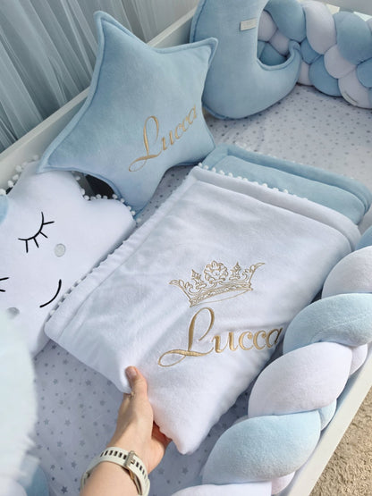 A baby bedding set in light blue and white, featuring a personalized blanket with a gold crown and the name 'Lucca', a matching star pillow, and a braided pillow, arranged in a baby crib. A hand is holding the blanket.