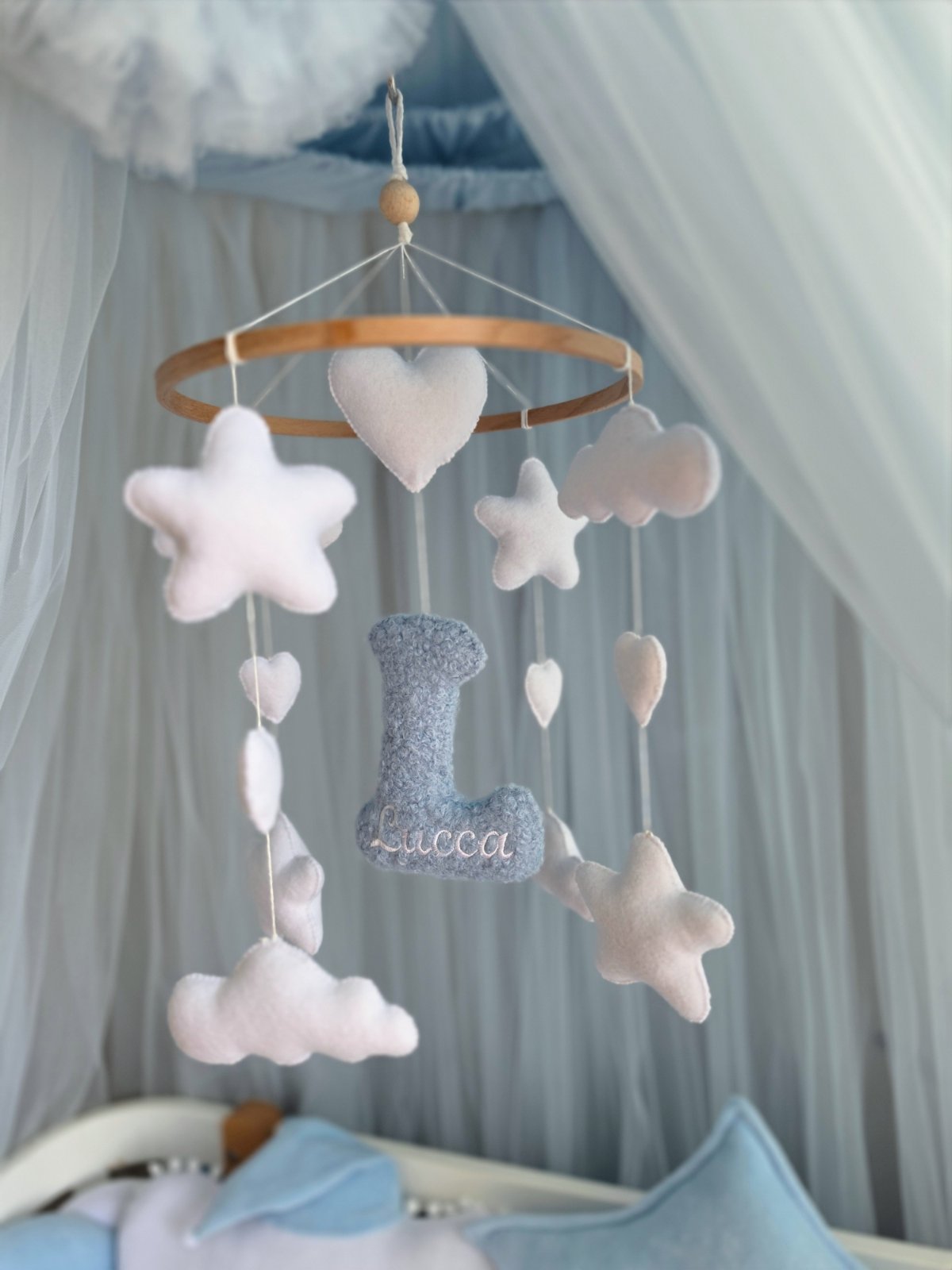 A baby nursery mobile hanging in front of a white canopy, featuring a light wood hoop with soft white felt stars, hearts, and clouds, plus a blue glittery felt letter 'L' with the name 'Lucca' embroidered in white.