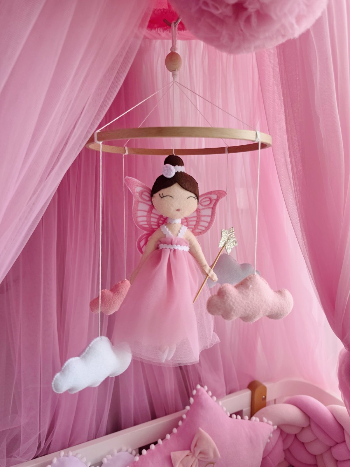 A pink fairy mobile hanging in a nursery crib, featuring a plush fairy doll in a pink dress with butterfly wings, surrounded by soft pink tulle canopy, clouds, and pillows.