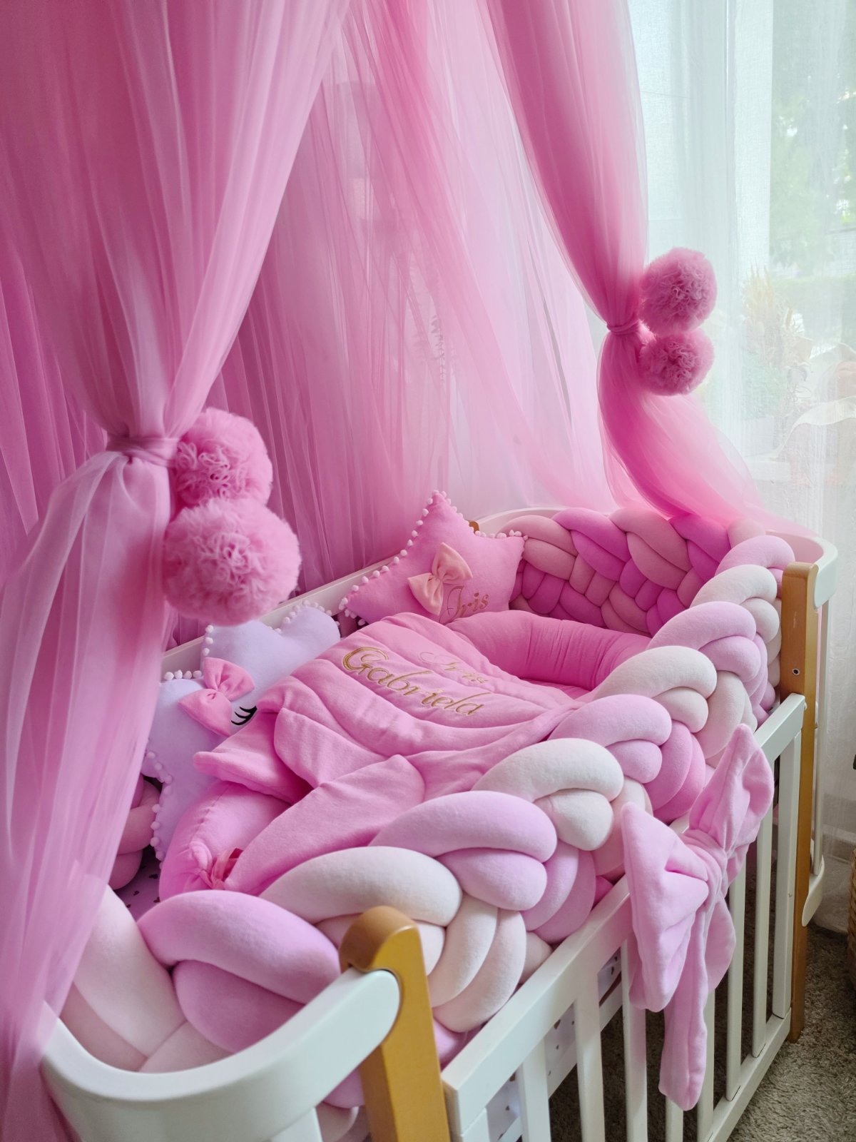 A pink baby crib setup with a chunky braided bumper pad, matching pink and white blankets, plush pillows, and a sheer pink canopy hanging over a white crib.