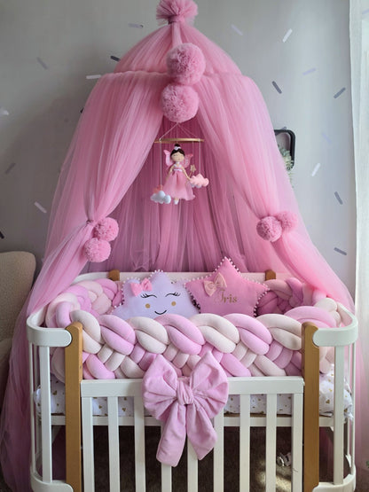 A pink baby canopy crib bedding set with a white wooden crib, chunky braided pads, a plush bow, and matching pillows under a sheer pink canopy with pom-poms.