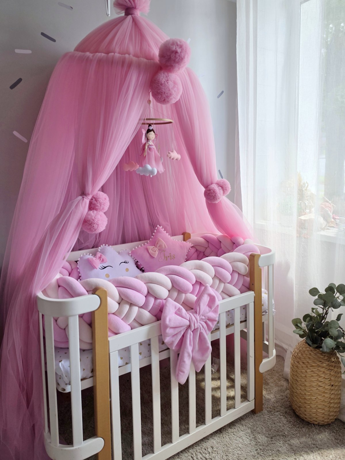 A pink baby canopy crib setup with a white wooden crib, pink tulle canopy, pink pom-poms, a hanging mobile, chunky pink and white chain pillow, and matching pink bedding in a softly lit nursery.