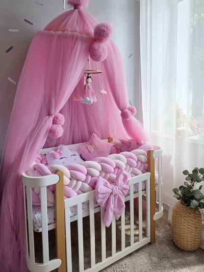 A pink baby canopy crib setup with a white wooden crib, pink tulle canopy, pink pom-poms, a hanging mobile, chunky pink and white chain pillow, and matching pink bedding in a softly lit nursery.