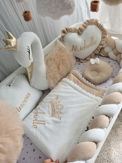 A styled baby bedding set in a nursery setting, featuring a white and beige plush swan pillow with a gold crown, a heart-shaped cushion embroidered with 'Michelle', a matching blanket with gold embroidery, and a braided pillow in white and beige.