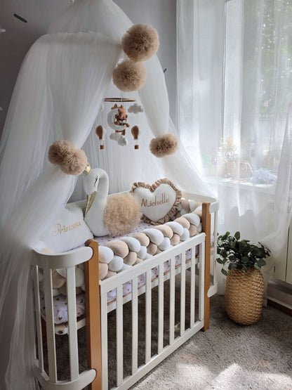 A styled baby nursery scene featuring a wooden crib with white bedding, a white goose plush, a heart-shaped pillow personalized with 'Michelle', a white canopy with fluffy pom-poms, and a hanging mobile, set in a softly lit room with sheer curtains and a potted plant.