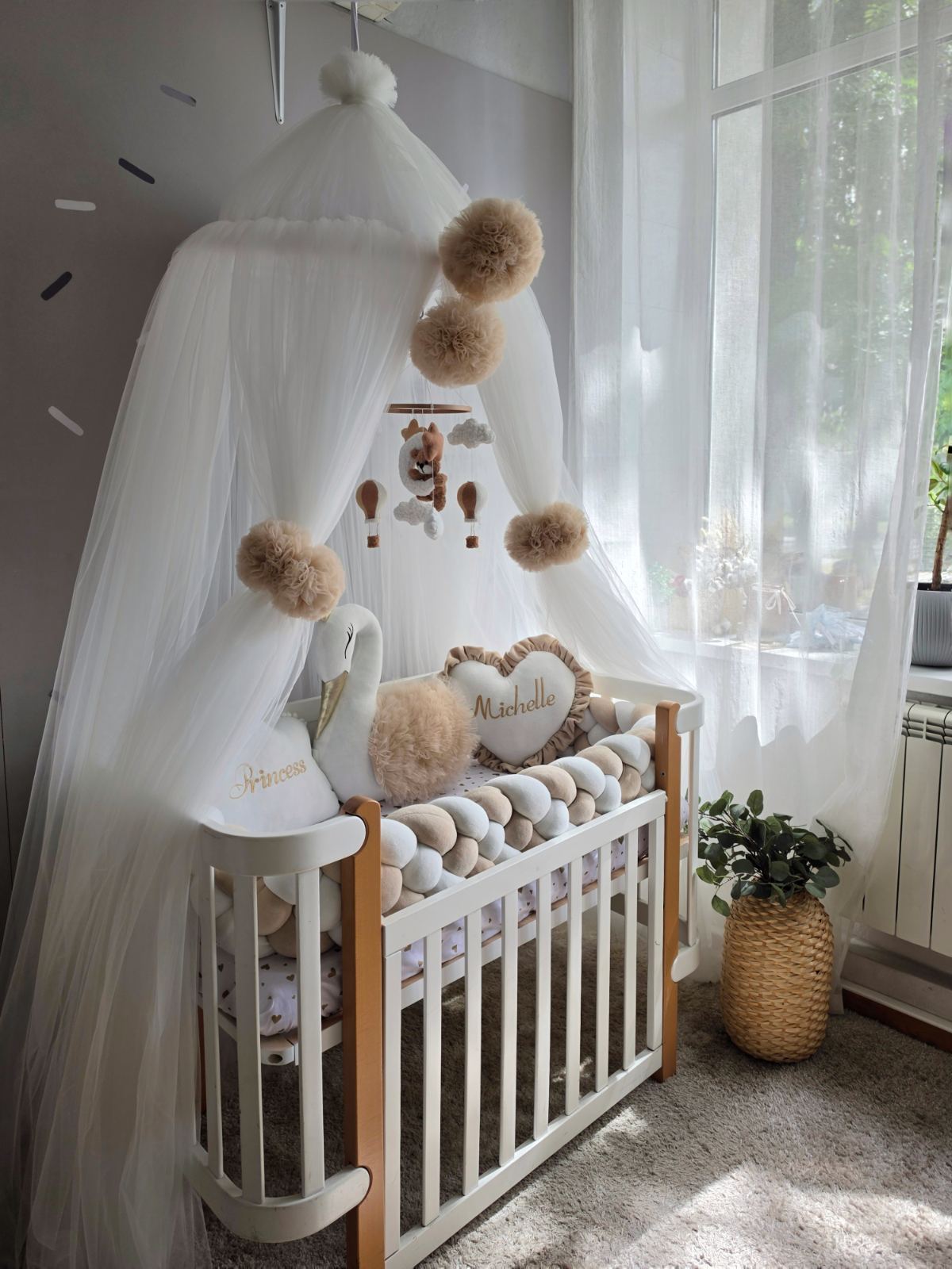 A styled baby nursery corner featuring a white wooden crib with a white canopy, beige pom-poms, a swan plush, and a heart-shaped pillow personalized with the name 'Michelle'.