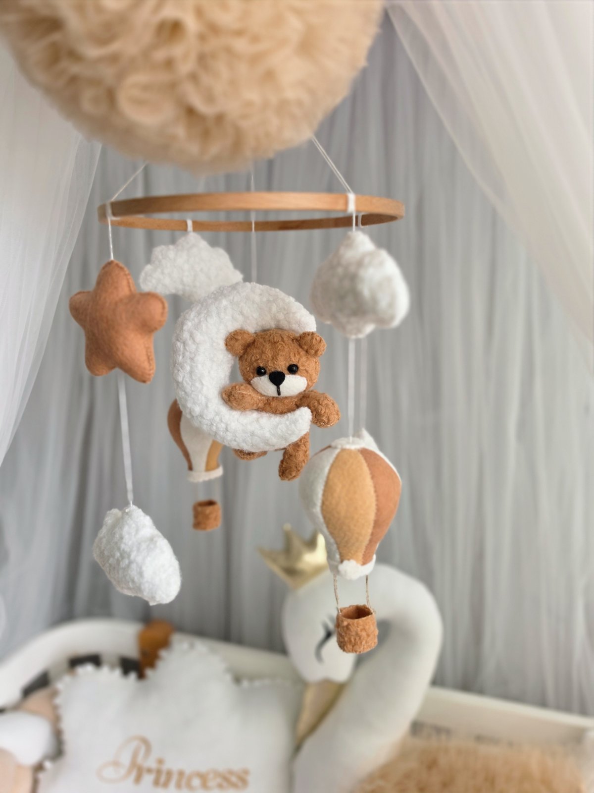 A soft baby mobile hanging in a nursery, featuring a teddy bear in a fluffy white crescent moon, white cloud shapes, a tan star, and a hot air balloon, with a wooden ring frame.