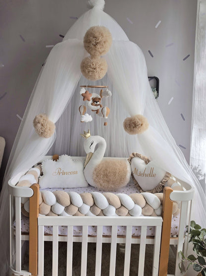 A white braided baby crib decor styled with a white canopy, beige pom-poms, a swan plush toy, and personalized pillows reading 'Princess' and 'Michelle' in gold script.