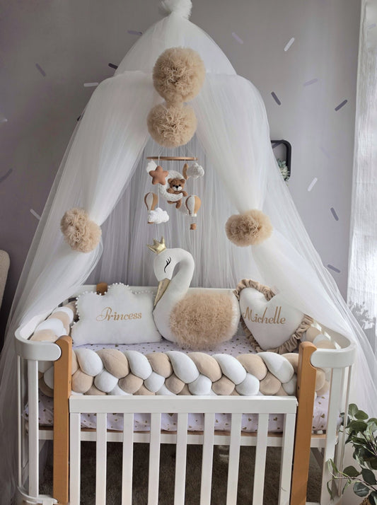 A white braided baby crib decor styled with a white canopy, beige pom-poms, a swan plush toy, and personalized pillows reading 'Princess' and 'Michelle' in gold script.