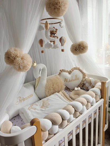 A styled baby nursery crib setup with a white wooden crib, cream and beige braid pillow, white canopy, plush swan toy, hot air balloon mobile, and a heart-shaped pillow embroidered with the name 'Michelle'.
