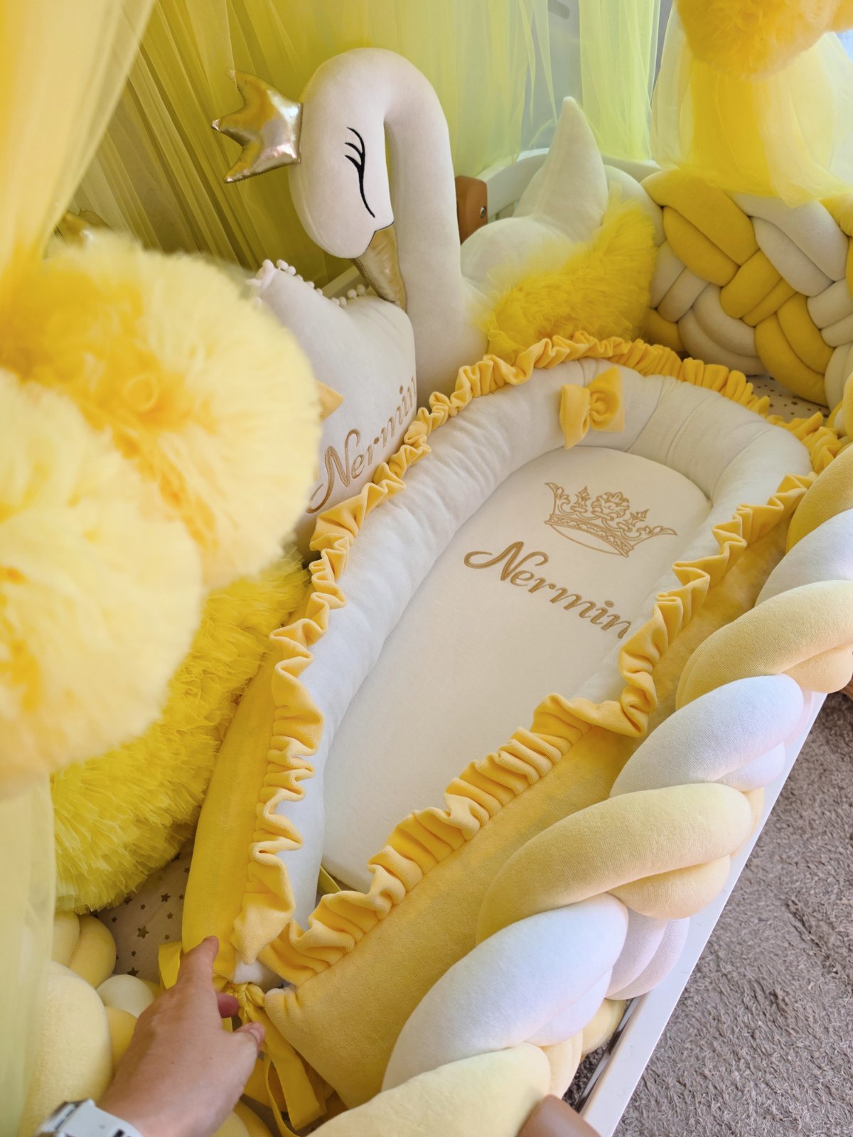 A yellow and white personalized baby Moses basket bassinet with ruffled trim, surrounded by plush yellow toys, a white swan plush, and a hand adjusting the bassinet fabric.