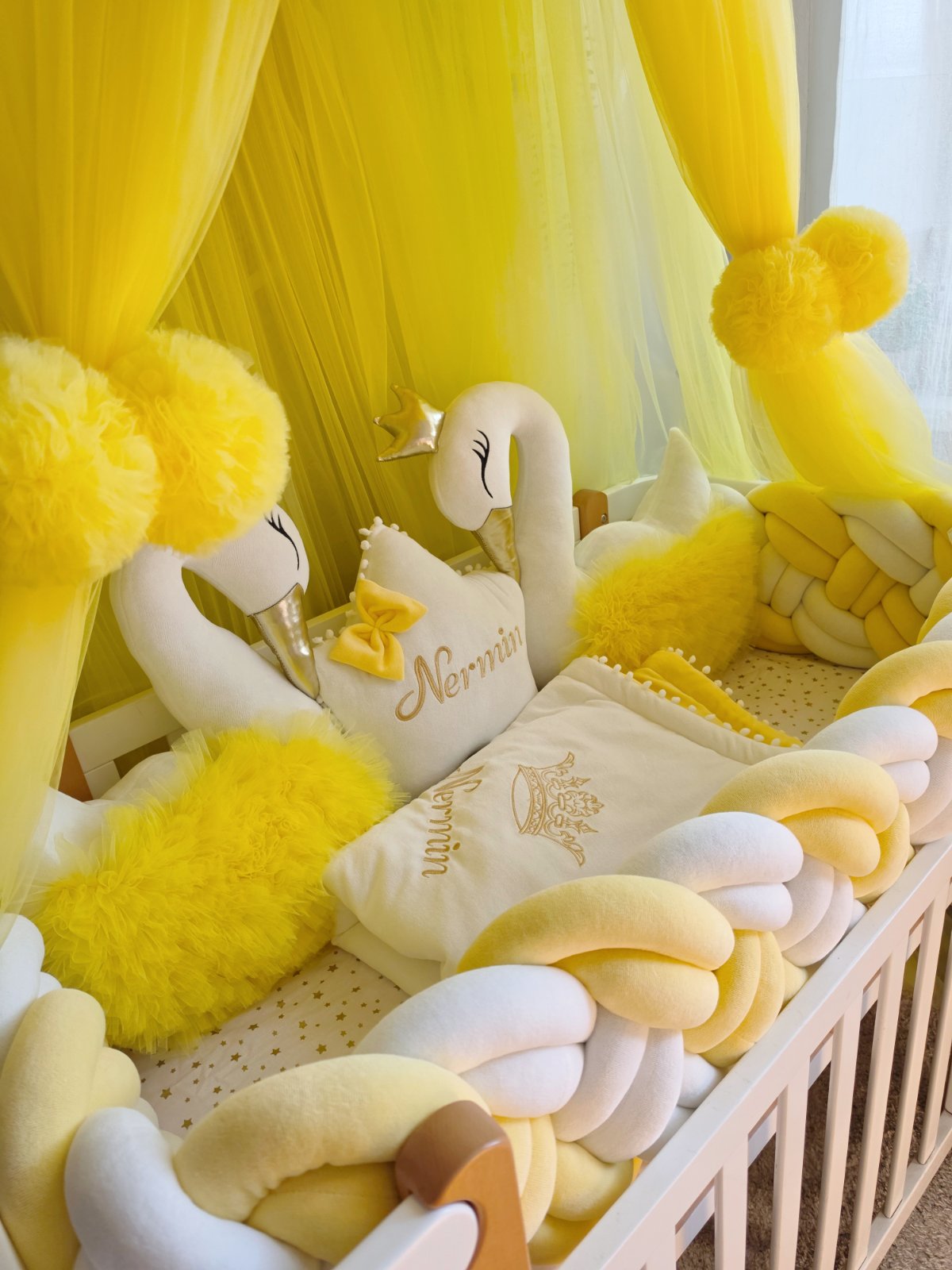 A yellow and cream baby crib set featuring a plush braided pillow, matching pillows, a white swan plush toy, and a yellow canopy with pom-poms.