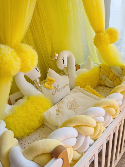 A yellow and cream baby crib set featuring a plush braided pillow, matching pillows, a white swan plush toy, and a yellow canopy with pom-poms.
