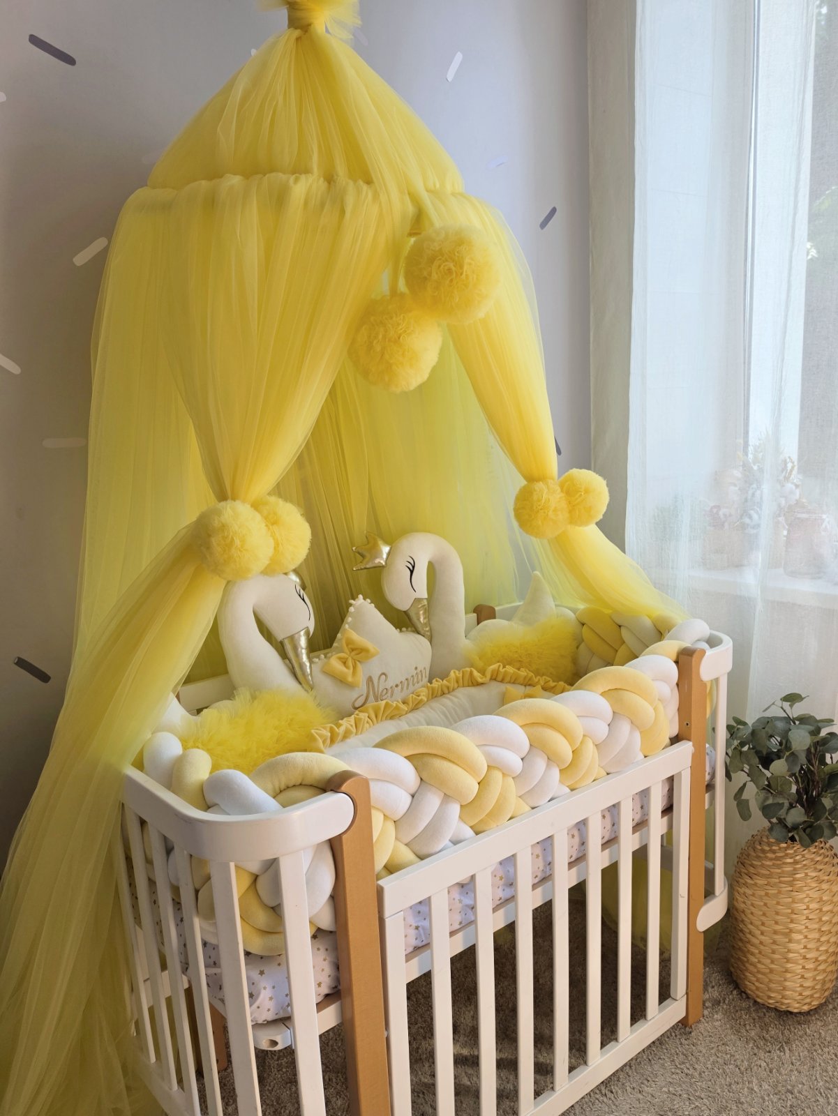 A yellow tulle baby canopy with fluffy pom-poms and matching yellow and white pillow snake guards draped over a white wooden crib in a softly lit nursery.