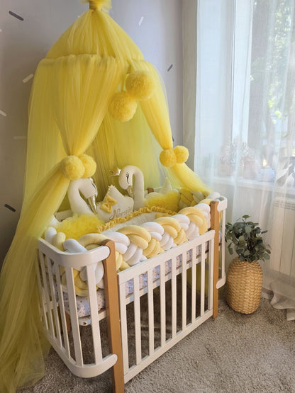 A white wooden baby crib styled with a bright yellow tulle canopy, yellow pom-poms, a plush swans, and yellow and white padded cushions in a softly lit nursery.