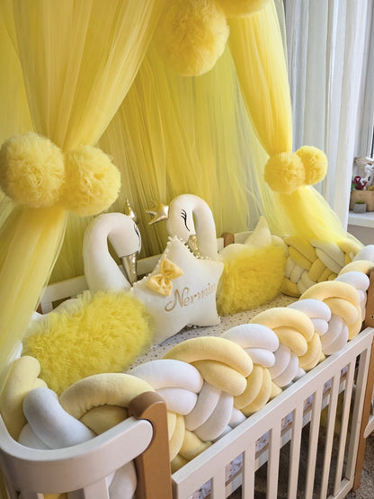 A yellow and white baby canopy crib setup with a sheer tulle canopy, fluffy pom-pom ties, a braided pad, two white swan plush toys, and a star pillow with the name 'Nora' on it.