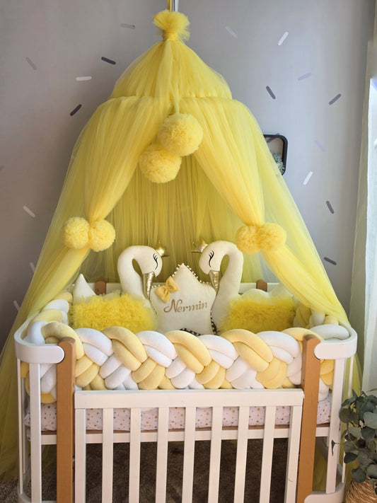 A yellow baby canopy with fluffy pom-poms draped over a white wooden crib, styled with yellow and white braided bedding, two white swan plush toys, and a central pillow that reads 'Nemmin'.